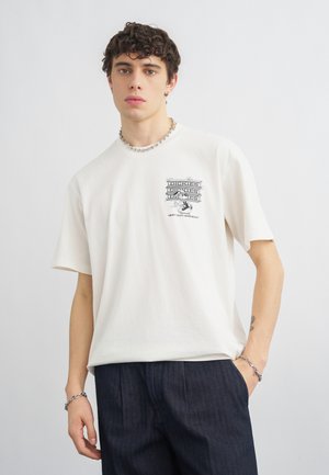 Mladý muž v bielom tričku Dickies s logom a tmavými nohavicami stojaci pred jednoduchým svetlým pozadím.