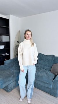 Krém színű garbós pulóver ribb szabású aljjal és mandzsettával, világoskék egyenes szárú farmernadrággal, oldalsó csíkkal. Kanapé és szörfdeszka háttér.