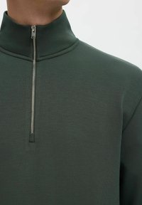 Felpa con zip corta di colore verde scuro, realizzata in tessuto liscio. Presenta un colletto alto e una zip argentata con un design semplice e moderno.