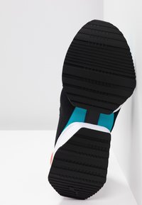 Semelle noire de chaussure de sport avec motif de bande de roulement texturé, montrant les parties supérieures bleues et blanches, placée contre un fond blanc et un mur.