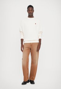 Mann iført off-white hettegenser og brune vide jeans som står mot en ensfarget hvit bakgrunn.