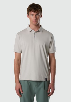 Joven con cabello rizado y corto, vistiendo una camisa polo de manga corta color beige claro y pantalones verdes, posando frente a un fondo liso.