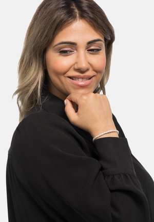 Silbernes Armband mit kleinen, glatten Perlen, das an einer Hand getragen wird, die nahe am Kinn ruht. Die Bluse ist schwarz mit gepufften Ärmeln.