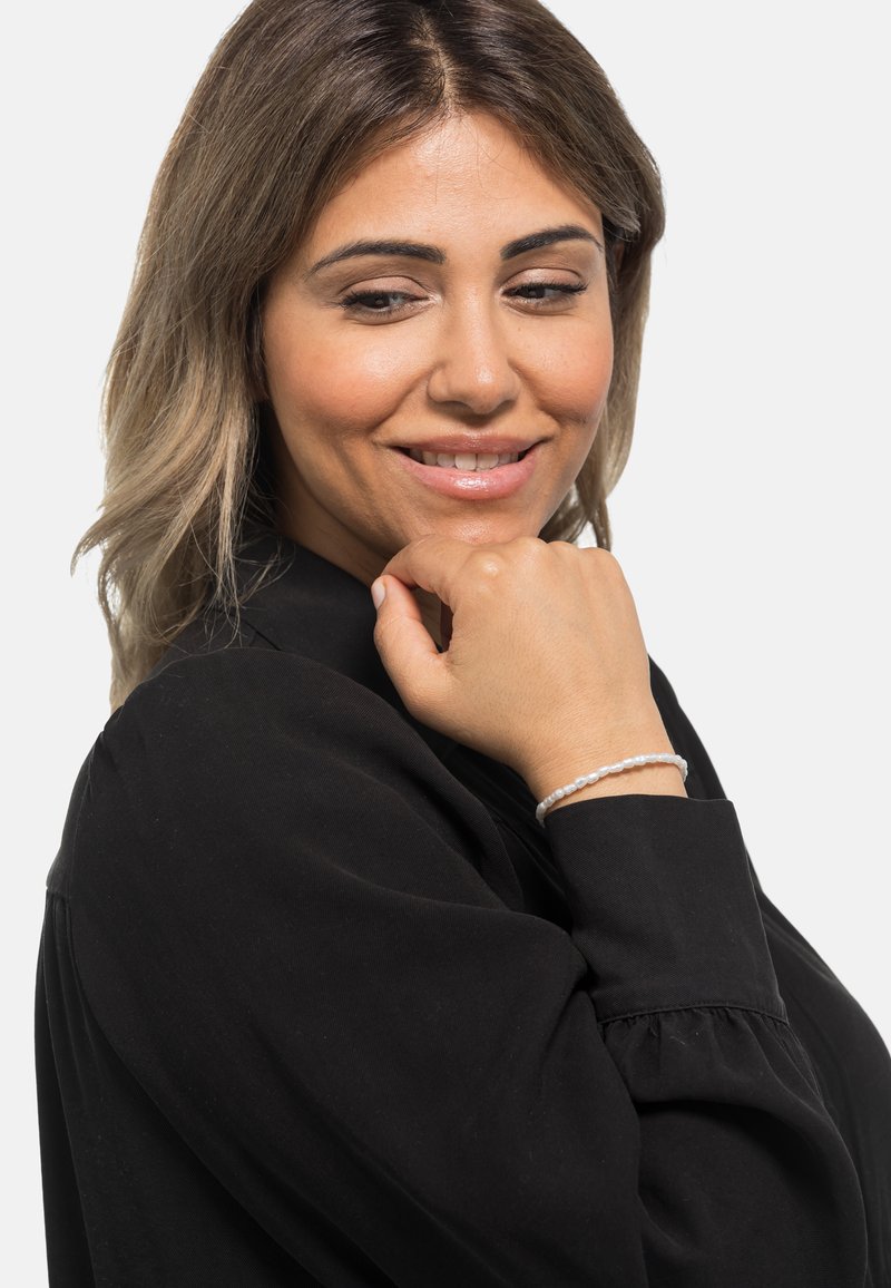 Bracciale in argento con piccole perline lisce indossato su una mano appoggiata vicino al mento. La blusa è nera con maniche a sbuffo.