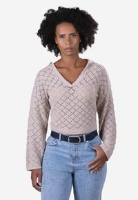 Ljust beige stickad tröja med V-ringning, diamantmönster och långa ärmar, tillsammans med högmidjade blå jeans och ett mörkt bälte.