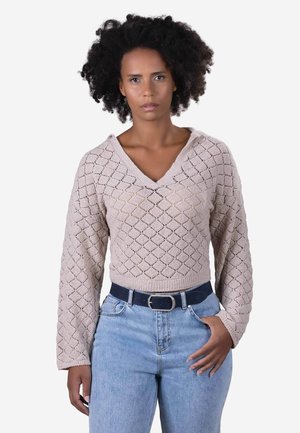 Suéter de punto beige claro con escote en V, patrón de rombos y mangas largas, combinado con jeans azules de cintura alta y un cinturón oscuro.