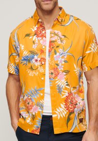 Chemise à manches courtes, à boutons, en tissu orange vif avec de grands motifs floraux en rose, bleu et vert, dotée d'un design de col classique.