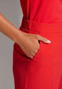 Rote Hose aus einem glatten, dehnbaren Stoff. Eine Nahaufnahme zeigt eine Hand in der Seitentasche und hebt das Design und die Passform der Tasche hervor.