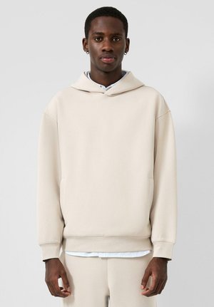 Jonge man die een beige hoodie en bijpassende broek draagt, staand tegen een effen lichtgrijze achtergrond.