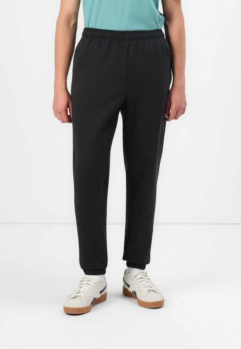 Μαύροι tapered jogger παντελόνες από μαλακό ύφασμα, με ελαστική μέση και μανσέτες στους αστραγάλους. Φοριούνται με λευκά αθλητικά παπούτσια.