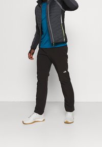 Veste isolante noire avec des accents gris et verts, chemise bleue, pantalon noir et baskets blanches sur un mannequin devant un fond neutre.