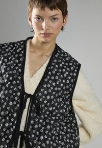 Czarna pikowana kamizelka z białymi wzorami kwiatowymi, z miękką kremową podszewką i czarnymi wstążkami z przodu.