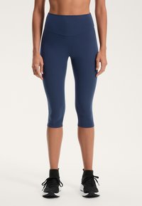 Temno modre capri legice iz gladkega, raztegljivega materiala; visok pas; skrajšana dolžina; obute s črnimi športnimi čevlji.