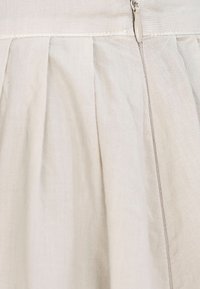 Faltenrock aus beigem, leichtem Stoff, mit einem verdeckten Reißverschluss und feinen Nähten entlang der Säume.