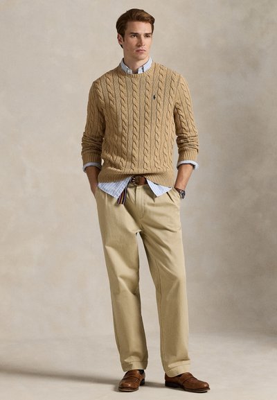 Jonge man met een beige kabelgebreide trui over een gestreept overhemd, beige broek, bruine leren riem, horloge en bruine loafers, staand met handen in de zakken.