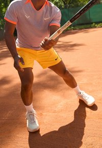 Kortärmad, ljuslila träningsskjorta med orange detaljer, gula shorts med svart logga och vita tennisskor på en grusplan.