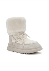 Bota de invierno en beige claro con forro de piel sintética y un eje acolchado. Cuenta con puntera redondeada, suela plana y un frente con cordones para ajuste.