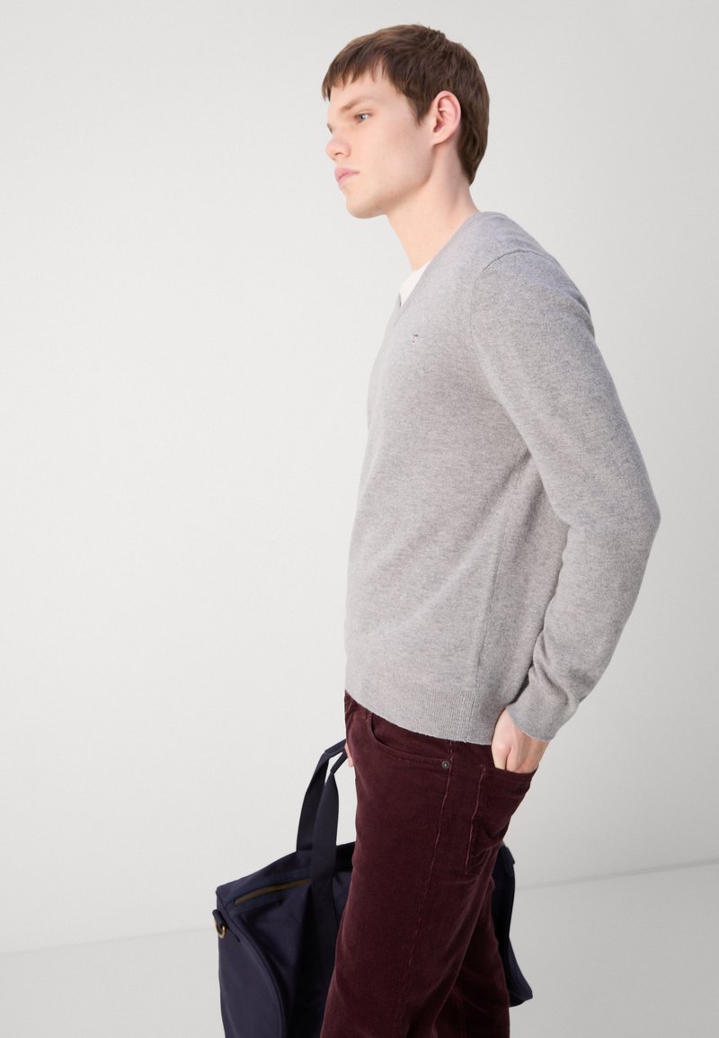 Grauer V-Ausschnitt-Pullover aus einem weichen Stoff, kombiniert mit burgunderfarbenen Cordhosen. Eine marineblaue Tasche wird in der Hand des Subjekts gehalten.