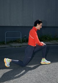 Rode sporttop met lange mouwen en hoge kraag, zwarte aansluitende leggings en meerkleurige hardloopschoenen. Buitensetting met een grijze achtergrond.