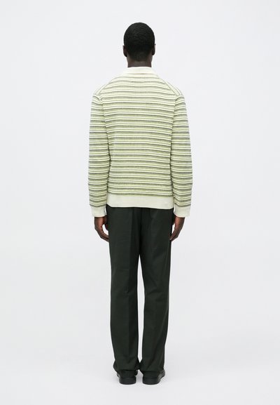 Man standing facing away wearing a cream and green striped sweater, black pants, and black shoes against a plain white background.