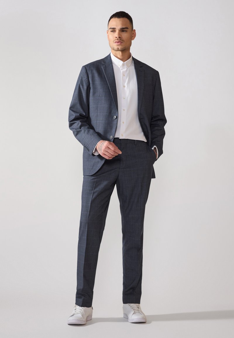 Completo checked blu scuro con giacca su misura e pantaloni slim-fit, abbinato a una camicia bianca e sneakers bianche.