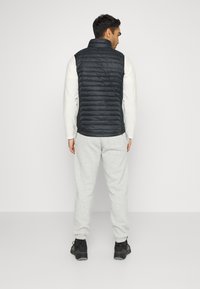 Gilet trapuntato nero con colletto alto, indossato sopra una camicia a maniche lunghe di colore chiaro, abbinato a pantaloni della tuta grigi e scarpe nere.
