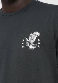 Juodas medvilninis marškinėliai su apvaliu iškirpimu, ant kairiojo krūtinės krašto pavaizduota balta grafika su ereliu ir penkiais žvaigždutėmis.