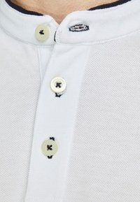 Nærbillede af farvet polo-skjorte krave i hvid med tre hvide knapper, hvoraf to er åbne, og sort syning omkring knaphullerne.