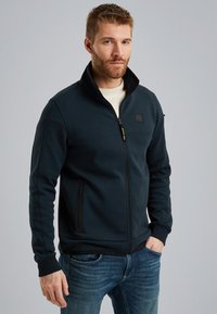 Donkerblauwe zip-up jas met hoge kraag, met ritssluiting zijzakken, geribbelde manchetten en een zachte stoftextuur. Draagt over een lichtgekleurde shirt.