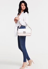 Borsa bianca con finitura liscia, accessori in oro e logo prominente. Esibita con una blusa bianca, jeans skinny blu e tacchi nudi.