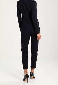 Mörkblå kostymjacka med smalrandigt mönster och åtsittande byxor med uppvikta ärmar, i kombination med svarta högklackade skor. Slät tygtextur.