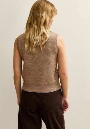 Blonde woman facing wall wearing a sleeveless brown knit vest and dark brown pants, standing with relaxed arms at sides.