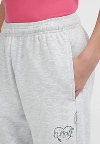 Persoon in lichte grijze sweatpants met elastische tailleband en hand in zak, roze top zichtbaar op de taille.