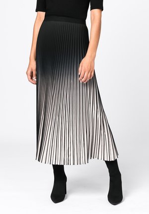 Nainen, joka pukeutunut mustavalkoiseen liukuvärjättyyn laskostettuun midi-hameeseen, yhdistettynä mustiin istuviin nilkkureihin, joissa on korkeat korot, valkoisella taustalla.