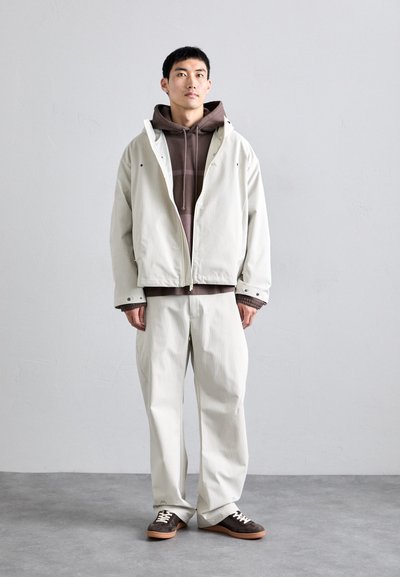 Conjunto casual de hombre que incluye una chaqueta beige clara, una sudadera marrón y pantalones en tonos neutros a juego. Zapatillas marrones con detalles blancos completan el look.