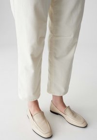 Mocassini in pelle beige con una superficie liscia; design slip-on con punta cucita e una suola bassa nera, indossati con pantaloni color crema.