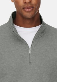Grå zip-up sweatshirt med høj krave og tekstureret stof. Har en sølv lynlås og et lagdelt hvidt skjorteunder.