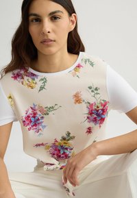 Blomstret kortærmet top med en cremefarvet krop, der har farverige blomstermønstre og en hvid halslinning. Stoffet har en glat tekstur.