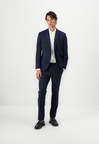 Costum bleumarin dintr-un material texturat, cu rever înclinat, asociat cu o cămașă albă. Loaferi negri completează ținuta. Design simplu, modern.
