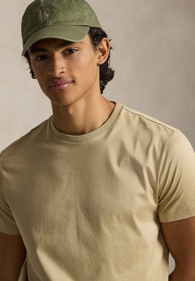 Ανοιχτό μπεζ κοντομάνικο μπλουζάκι με στρογγυλή λαιμόκοψη, συνδυασμένο με σκούρο πράσινο καπέλο μπέιζμπολ με λογότυπο. Ομαλή υφή υφάσματος.