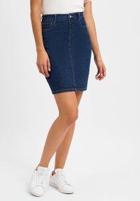 Dunkelblaue Jeansrock mit tailliertem Schnitt, knielang, mit vorderen Taschen und Kontrastnähten. Kombiniert mit weißen Sneakern.