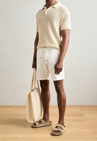 Polo shirt beige in maglia, pantaloni corti bianchi con coulisse, sandali scivolati grigi e una borsa beige. Sfondo in legno.