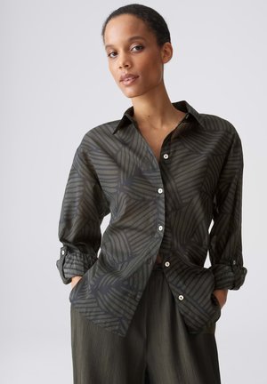 Femme portant une chemise boutonnée à motif vert foncé avec manches retroussées et pantalon ample assorti, posant les mains dans les poches devant un fond uni.