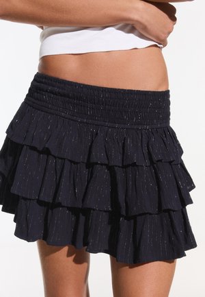 Kobieta w czarnej, wielowarstwowej spódnicy z falbanami z teksturowanej tkaniny z subtelnymi, błyszczącymi paskami, zestawionej z białym, krótkim topem.