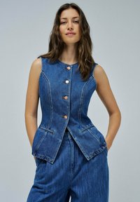 Gilet en denim à lavage bleu foncé, avec cinq boutons en cuivre, coupe structurée, sans manches, et deux poches frontales avec des coutures contrastées.