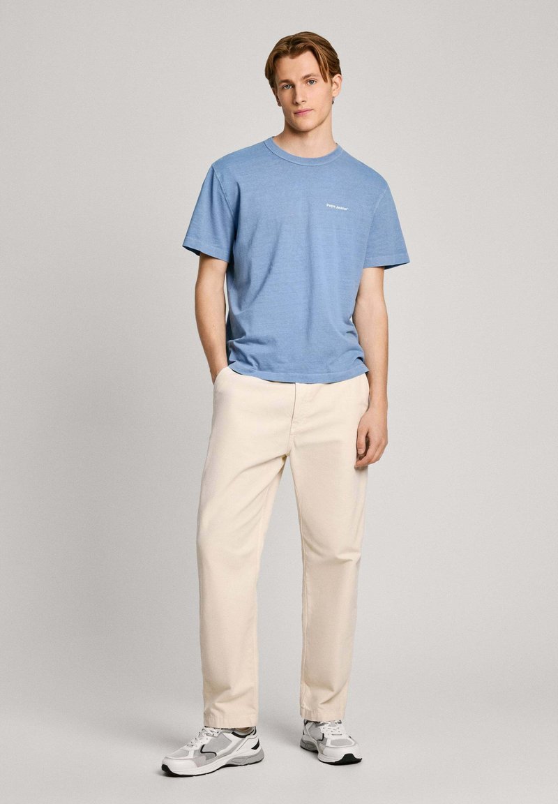 Himmelblaues T-Shirt mit kurzen Ärmeln und Rundhalsausschnitt, mit weißem Logo auf der Brust, kombiniert mit cremefarbenen Hosen und grau-weißen Sneakers.