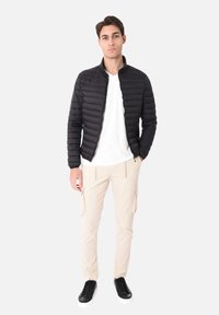 Giacca leggera nera imbottita con colletto rialzato, abbinata a una maglietta bianca e pantaloni cargo beige. Sneakers nere completano l'outfit.