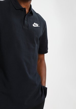 Homme portant un polo noir Nike avec un logo Swoosh blanc, debout avec une main dans la poche devant un fond gris clair uni.