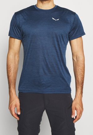 Mies, jolla on istuva, lyhythihainen tummansininen t-paita, jossa vasemman rinnan kohdalla pieni valkoinen logo, sekä mustat housut.