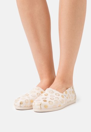 Baskets en toile à enfiler avec une base beige clair, décorées de motifs de soleil doré et de fleurs. Bout arrondi et semelle légèrement texturée.